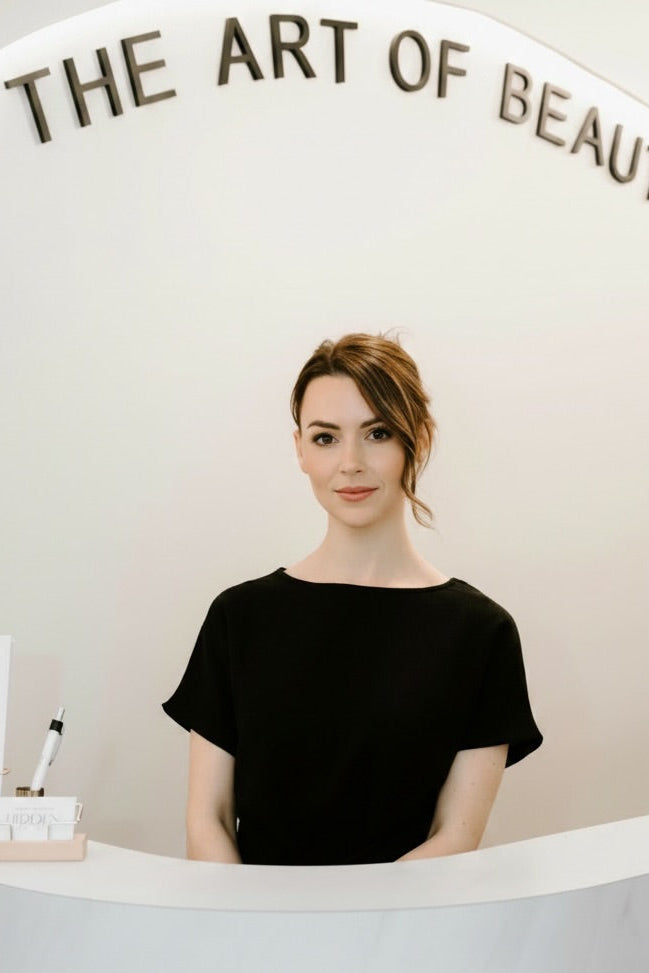 A woman with styled brown hair stands behind a white counter in a bright, minimalist interior. She is wearing a black short-sleeve top.